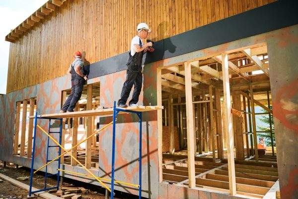 Carpenters constructing wooden framed house. Two men workers cladding facade of house with cement particle boards, fastening them with screwdrivers. Concept of modern eco-friendly construction.